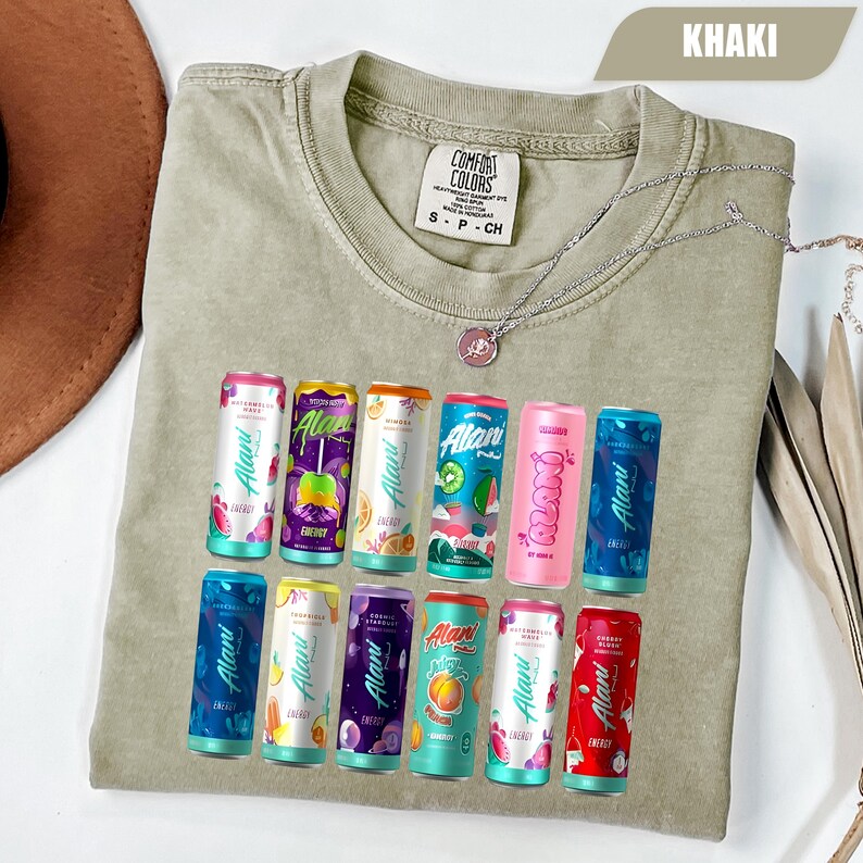 May include: A khaki-colored crew neck t-shirt featuring a graphic print of twelve colorful Alani Nu energy drink cans arranged in two rows. The shirt has a Comfort Colors brand label at the inner neckline and a small silver-toned pendant necklace.