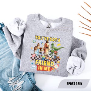 May include: A heather grey crewneck sweatshirt featuring a graphic print of a central figure walking with various cartoon characters. The text reads YOU'VE GOT A FRIEND IN ME. The design includes planets, stars, and a rocket ship on a checkered base.