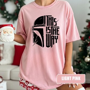 May include: Light pink t-shirt featuring a black graphic of a helmet and the text "THIS IS THE WAY." The shirt is a solid light pink color. The words "LIGHT PINK" are in a rectangular box at the bottom of the shirt.