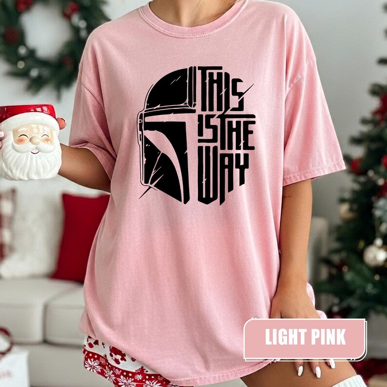 May include: Light pink t-shirt featuring a black graphic of a helmet and the text "THIS IS THE WAY." The shirt is a solid light pink color. The words "LIGHT PINK" are in a rectangular box at the bottom of the shirt.
