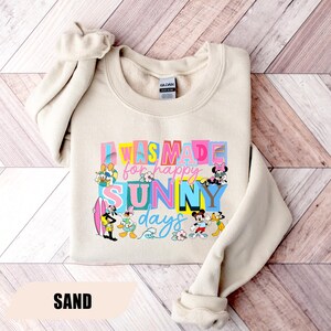May include: Sand-colored sweatshirt with the text "I WAS MADE for happy SUNNY days" in colorful block letters. Disney characters like Mickey Mouse, Donald Duck, and Minnie Mouse are featured in the design. The sweatshirt has a crew neck and long sleeves.