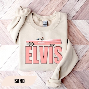 May include: Sand-colored sweatshirt featuring a pink vintage car graphic with the text "ELVIS" in large pink letters. The car has a person driving it. The word "SAND" is printed at the bottom of the sweatshirt.