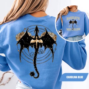 May include: Carolina blue sweatshirt featuring a black dragon design with gold accents on the back. The dragon is surrounded by a constellation and cloud design. The front of the sweatshirt has a smaller image of the dragon and the text "BASGIATH WAR COLLEGE DRAGON RIDERS".