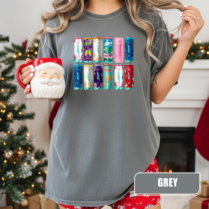 May include: A charcoal grey t-shirt featuring a graphic print of twelve colorful Alani Nu energy drink cans arranged in two rows. The shirt is paired with a white ceramic Santa Claus mug with a red handle and a red rim, held in one hand.
