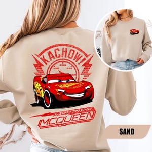 May include: A sand-colored crewneck sweatshirt featuring a large graphic on the back of the red race car Lightning McQueen with the text KACHOW and LIGHTNING MCQUEEN. A smaller version of the red race car is printed on the front left chest area.