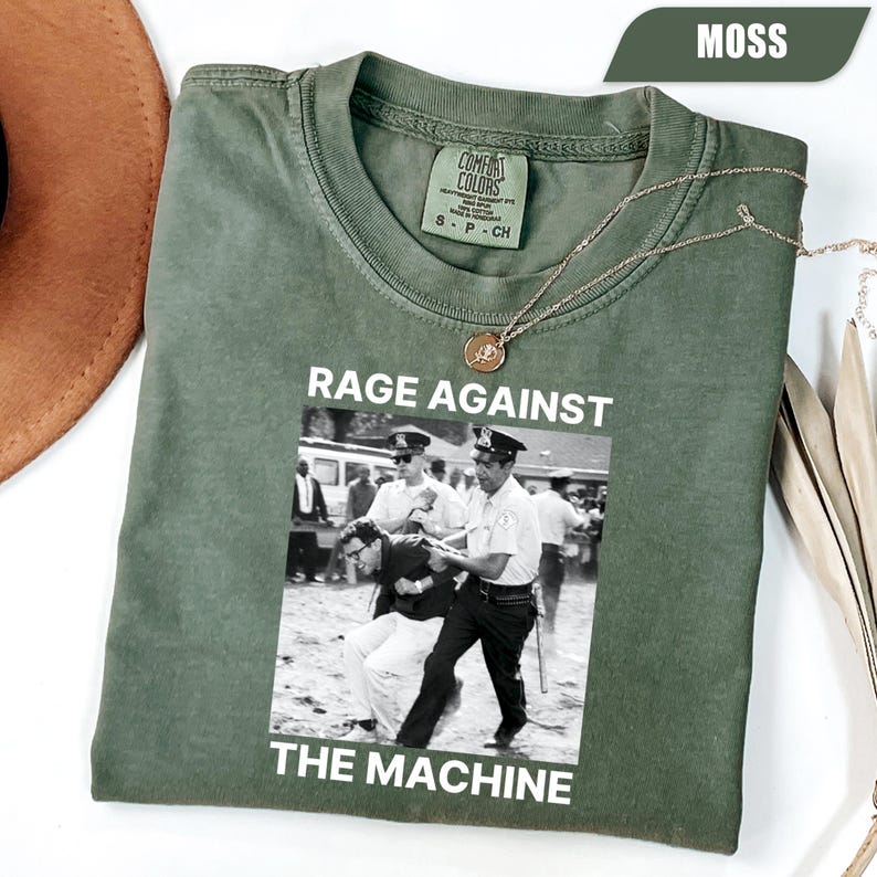 May include: A moss green t-shirt with a black and white graphic depicting a protest scene. The text "RAGE AGAINST THE MACHINE" is printed above and below the image. A gold necklace with a pendant is draped on the shirt.