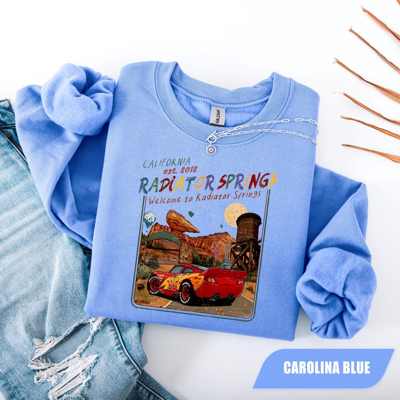 May include: A Carolina blue crewneck sweatshirt featuring a graphic print of a red race car on a desert road. The text reads California est. 2012 RADIATOR SPRINGS Welcome to Radiator Springs. The design includes a rock formation and water tower.