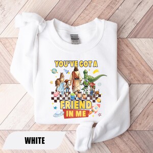 May include: A white long-sleeve sweatshirt featuring a graphic print of a central figure holding hands with various animated characters. The text "YOU'VE GOT A FRIEND IN ME" is printed in yellow and red. The design includes planets, stars, and a rocket.