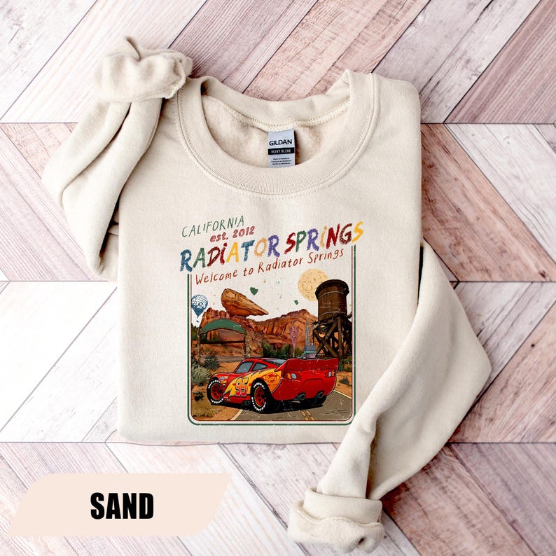 May include: A sand-coloured crewneck sweatshirt featuring a graphic print of a red race car on a desert road. The text reads California est. 2012 RADIATOR SPRINGS Welcome to Radiator Springs, with illustrations of a rock formation and water tower.