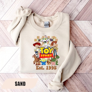 Puede incluir: Sudadera color arena con un gr&aacute;fico colorido de los personajes de Toy Story, incluyendo a Woody, Buzz Lightyear y Jessie. Las palabras "TOY STORY" est&aacute;n en letras grandes y en negrita, con "Est. 1995" debajo. La palabra "SAND" est&aacute; en la parte inferior.