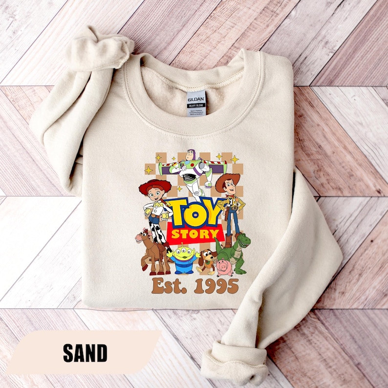 Puede incluir: Sudadera color arena con un gr&aacute;fico colorido de los personajes de Toy Story, incluyendo a Woody, Buzz Lightyear y Jessie. Las palabras "TOY STORY" est&aacute;n en letras grandes y en negrita, con "Est. 1995" debajo. La palabra "SAND" est&aacute; en la parte inferior.