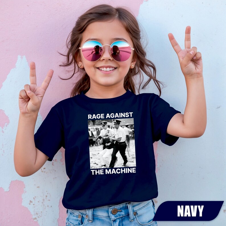 May include: Navy blue t-shirt featuring a black and white image and the text "RAGE AGAINST THE MACHINE". The shirt has a crew neck and short sleeves. The word "NAVY" is at the bottom of the shirt.
