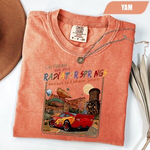 May include: A yam-colored short-sleeve t-shirt featuring a graphic print of a red race car with the number 95, a desert landscape, and the text "California est. 2012 RADIATOR SPRINGS Welcome to Radiator Springs" inside a rectangular frame.
