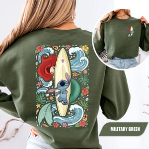 May include: A military green crewneck sweatshirt featuring a large graphic on the back of Stitch and Ariel from The Little Mermaid with a surfboard, ocean waves, tropical flowers, and ukuleles. A smaller version of the graphic is on the front.