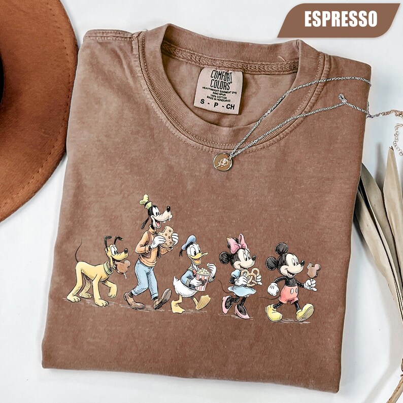 May include: An espresso-colored crew neck t-shirt featuring a graphic print of Pluto, Goofy, Donald Duck, Minnie Mouse, and Mickey Mouse walking together. The shirt has a Comfort Colors brand label at the inner neckline and a silver-toned necklace.