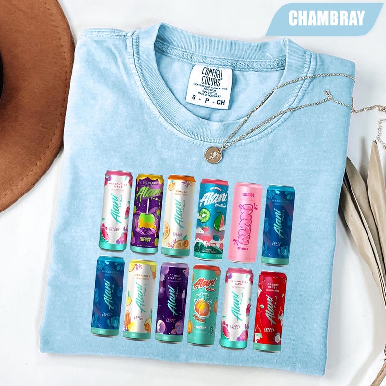 May include: A light blue t-shirt with a graphic print featuring twelve colorful Alani energy drink cans arranged in two rows. The shirt has a crew neckline, a Comfort Colors brand label, and a thin silver-toned chain necklace resting on top.