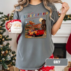 May include: A grey short-sleeve t-shirt featuring a graphic of a red race car in a desert landscape with the text "California Est. 2012 Radiator Springs" and "Welcome to Radiator Springs" printed above the car, held alongside a Santa Claus mug.