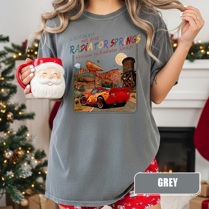 May include: A grey short-sleeve t-shirt featuring a graphic of a red race car in a desert landscape with the text "California Est. 2012 Radiator Springs" and "Welcome to Radiator Springs" printed above the car, held alongside a Santa Claus mug.