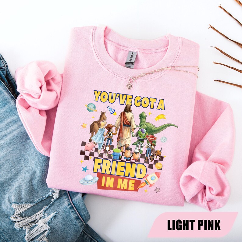 May include: A light pink crewneck sweatshirt featuring a graphic print of a central figure walking with various animated toy characters. The text reads "YOU'VE GOT A FRIEND IN ME" in yellow letters. The design includes planets, rockets, and a checkered floor.
