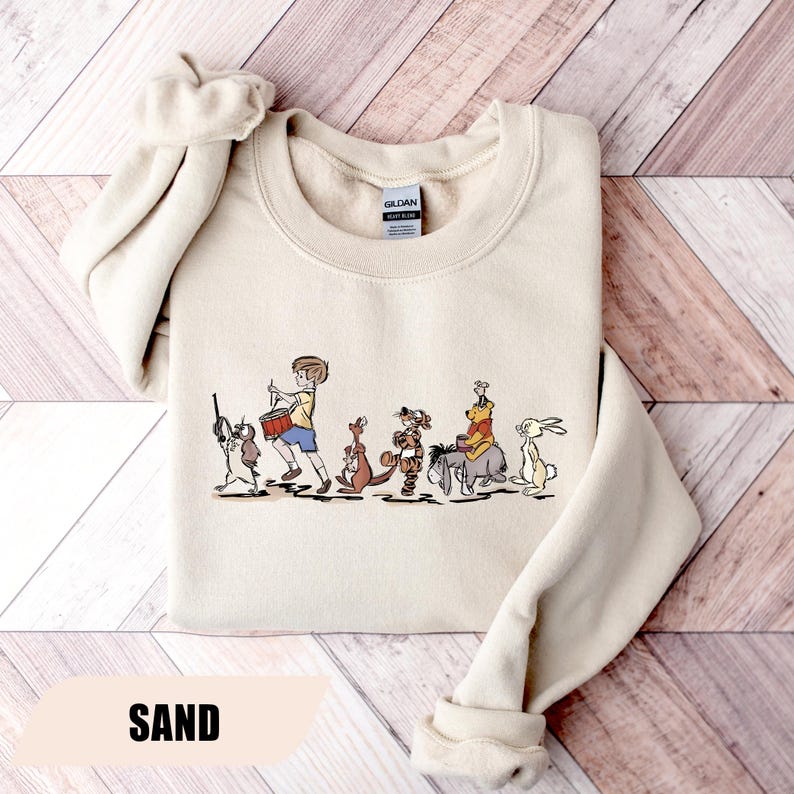 May include: Sand-colored sweatshirt featuring a line of cartoon characters, including a boy with a drum, a kangaroo, a tiger, Winnie the Pooh on a donkey, and a rabbit. The word "SAND" is printed at the bottom.