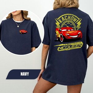 May include: A navy blue t-shirt featuring a small graphic of the red race car Lightning McQueen on the front left chest. The back displays a large yellow graphic with the text KACHOW and LIGHTNING MCQUEEN surrounding the red race car.
