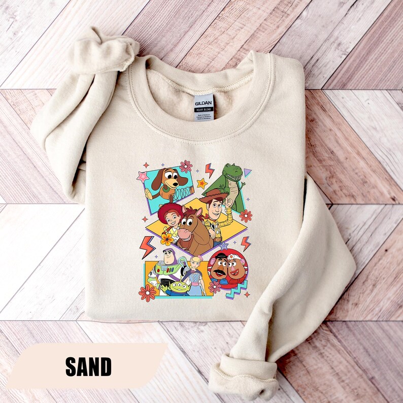 Puede incluir: Sudadera color arena con un gr&aacute;fico colorido de personajes de una pel&iacute;cula animada. El dise&ntilde;o incluye un perro, un dinosaurio, un vaquero y otros personajes. La palabra "SAND" est&aacute; impresa en la parte inferior de la sudadera.