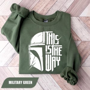 May include: Military green sweatshirt with a white graphic of a helmet and the text "THIS IS THE WAY". The sweatshirt is folded, showing the sleeves and the crew neck. The words "MILITARY GREEN" are in a banner at the bottom.