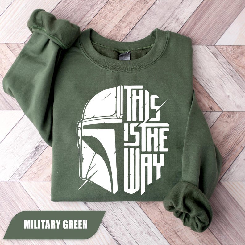 May include: Military green sweatshirt with a white graphic of a helmet and the text "THIS IS THE WAY". The sweatshirt is folded, showing the sleeves and the crew neck. The words "MILITARY GREEN" are in a banner at the bottom.