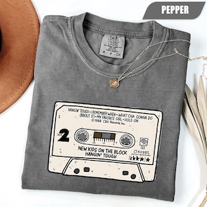 Op de afbeelding: Pepperkleurig T-shirt met een cassettebandje-afbeelding met de tekst "New Kids on the Block Hangin' Tough". Het shirt bevat songtitels en het label van het merk Comfort Colors. Een gouden ketting is over het shirt gedrapeerd.