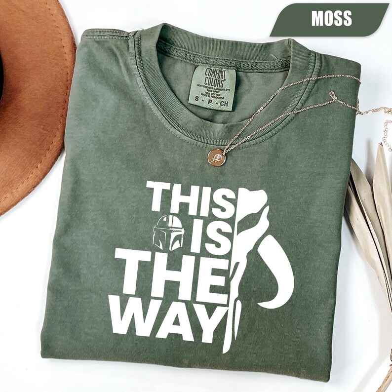 May include: A moss green short-sleeve t-shirt featuring the white text "THIS IS THE WAY" alongside a graphic of a helmet. The shirt has a crew neckline and a Comfort Colors brand label on the inside collar. The fabric has a washed, vintage appearance.