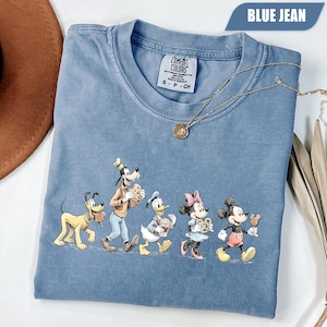 May include: A blue jean colored short-sleeve t-shirt featuring a graphic of Pluto, Goofy, Donald Duck, Minnie Mouse, and Mickey Mouse eating snacks. The shirt has a Comfort Colors label at the neck and a gold-toned chain necklace resting on it.