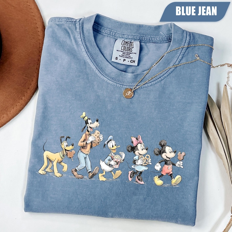 May include: A blue jean colored short-sleeve t-shirt featuring a graphic of Pluto, Goofy, Donald Duck, Minnie Mouse, and Mickey Mouse eating snacks. The shirt has a Comfort Colors label at the neck and a gold-toned chain necklace resting on it.