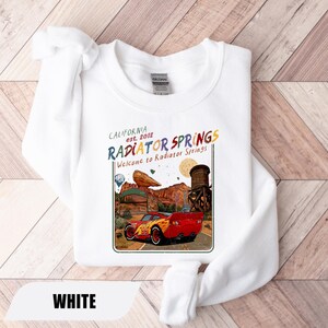 May include: A white crewneck sweatshirt featuring a graphic print of a red race car with the number 95 on the side. The design includes the text California est. 2012 Radiator Springs and Welcome to Radiator Springs, depicting a desert landscape.
