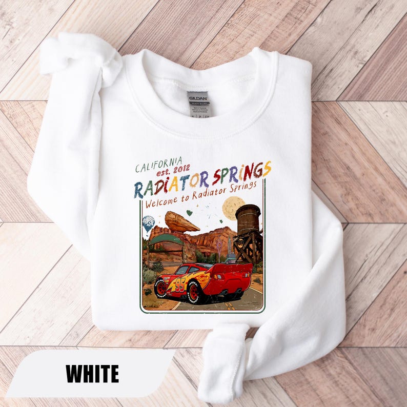 May include: A white crewneck sweatshirt featuring a graphic print of a red race car with the number 95 on the side. The design includes the text California est. 2012 Radiator Springs and Welcome to Radiator Springs, depicting a desert landscape.