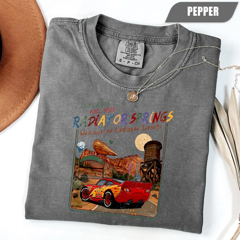 May include: A charcoal grey t-shirt featuring a graphic of a red race car with the number 95, set against a desert landscape with a water tower and rock formations. The text reads California Est. 2006 Radiator Springs Welcome to Radiator Springs.