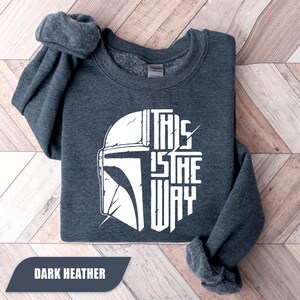 May include: Dark heather gray sweatshirt with a white graphic of a helmet and the text "THIS IS THE WAY". The sweatshirt is folded neatly on a wooden surface. The text "DARK HEATHER" is in a dark gray rectangle at the bottom.