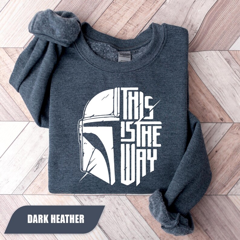 May include: Dark heather gray sweatshirt with a white graphic of a helmet and the text "THIS IS THE WAY". The sweatshirt is folded neatly on a wooden surface. The text "DARK HEATHER" is in a dark gray rectangle at the bottom.