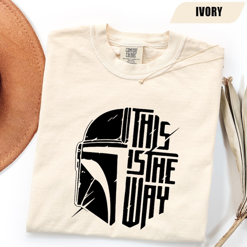 May include: Ivory-colored t-shirt with a black graphic of a helmet and the text "THIS IS THE WAY." The shirt is made of a soft material and has a crew neck.