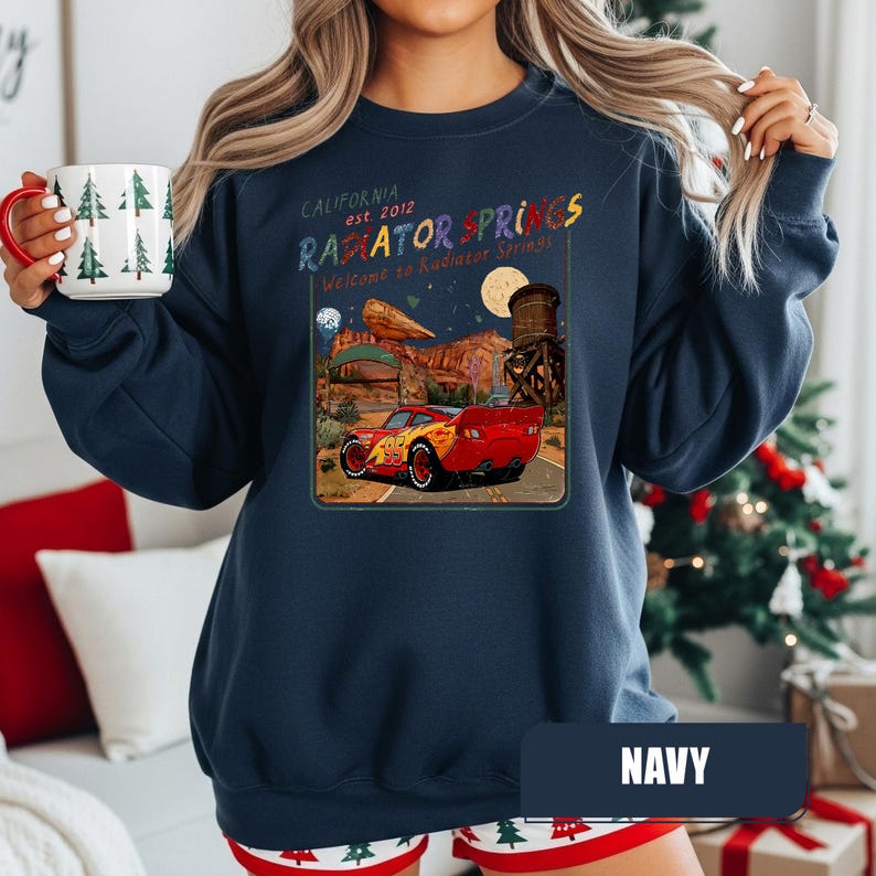 May include: A navy blue crewneck sweatshirt featuring a graphic print of a red race car on a desert road. The text reads California est. 2012 Radiator Springs Welcome to Radiator Springs, accompanied by a desert landscape, a water tower, and a full moon.
