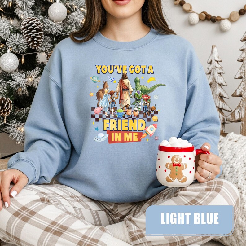 May include: A light blue crewneck sweatshirt featuring a graphic print of various cartoon characters walking with a central figure. The text reads "YOU'VE GOT A FRIEND IN ME" in yellow and red. A white ceramic mug with a gingerbread man design is also shown.