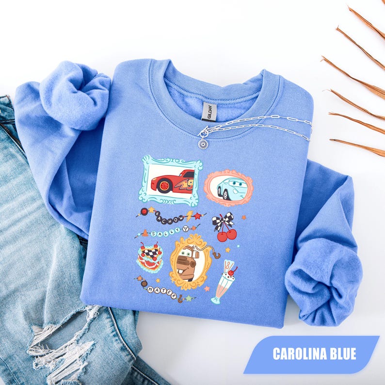 Puede incluir: Una sudadera de cuello redondo en color azul Carolina con un estampado gr&aacute;fico de personajes de la pel&iacute;cula Cars, incluyendo a Rayo McQueen, Sally y Mate en marcos ornamentados, junto a ilustraciones de cerezas, un batido y cuentas con letras.
