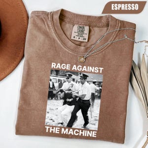 May include: Espresso-colored t-shirt with a black and white image of a protest scene. The text "RAGE AGAINST THE MACHINE" is printed in white. The shirt has a crew neck and short sleeves. A silver necklace with a pendant is draped across the shirt.