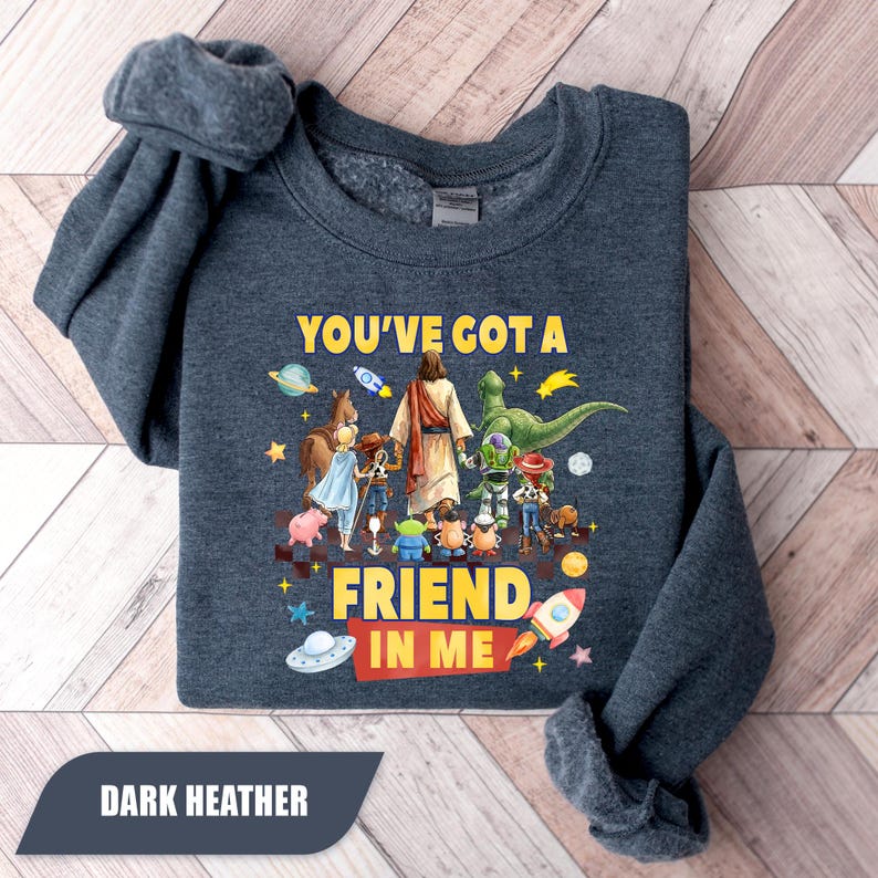 May include: A dark heather gray crewneck sweatshirt featuring a graphic print of a central figure in a robe surrounded by various animated toy characters. The text "YOU'VE GOT A FRIEND IN ME" is printed in yellow and white with space-themed illustrations.