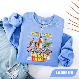 May include: A light blue crewneck sweatshirt featuring a graphic print of a central figure walking with various cartoon characters. The text "YOU'VE GOT A FRIEND IN ME" is printed in yellow and red. The design includes space-themed elements and a checkered pattern.