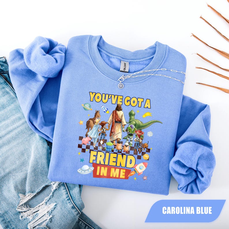 May include: A light blue crewneck sweatshirt featuring a graphic print of a central figure walking with various cartoon characters. The text "YOU'VE GOT A FRIEND IN ME" is printed in yellow and red. The design includes space-themed elements and a checkered pattern.