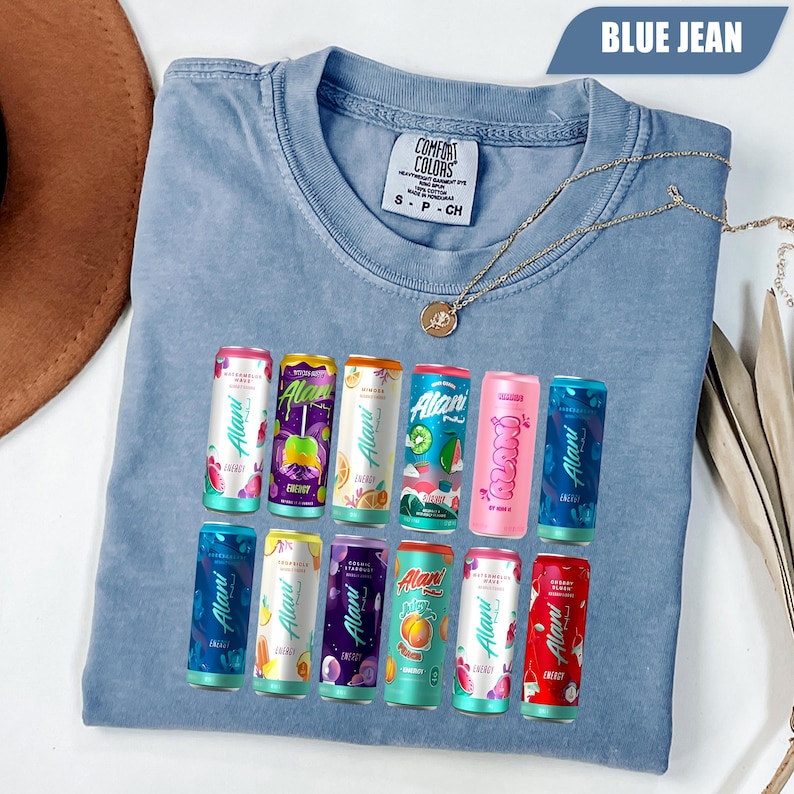 May include: A blue jean colored t-shirt with a Comfort Colors label at the neck. The front features a graphic print of twelve colorful Alani energy drink cans arranged in two rows, showcasing various fruit flavors and branding text on each can.