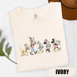 May include: An ivory crew neck t-shirt featuring a graphic print of Pluto, Goofy, Donald Duck, Minnie Mouse, and Mickey Mouse walking in a line. The characters are illustrated in a sketch style, and the shirt has a Comfort Colors brand label.