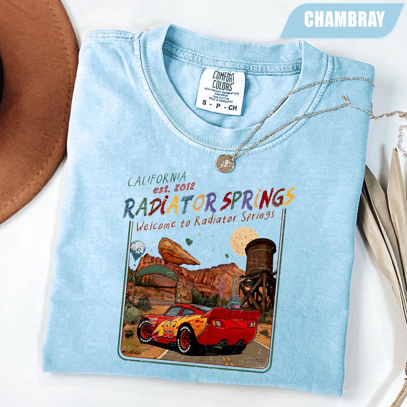May include: A light blue t-shirt featuring a graphic print of a red race car with the number 95, a desert landscape, and a water tower. The text reads California est. 2012 Radiator Springs Welcome to Radiator Springs. The shirt has a Comfort Colors label.