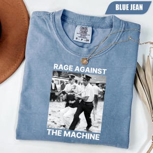 May include: Blue jean-colored t-shirt with the text "RAGE AGAINST THE MACHINE" and a black and white image of a protest. The shirt has a crew neck and short sleeves. A gold necklace is draped across the shirt.