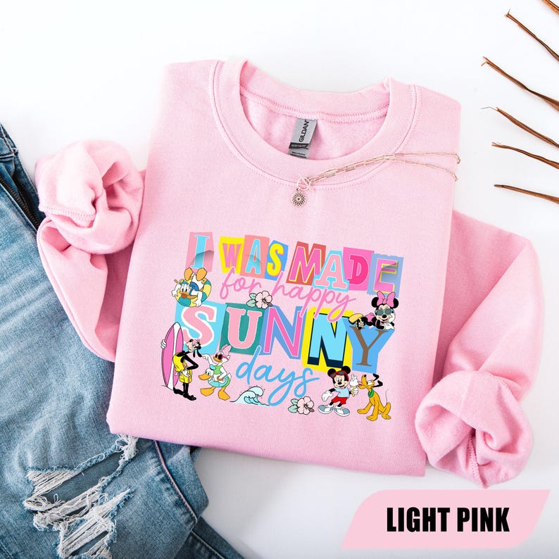 May include: Light pink sweatshirt with the text "I WAS MADE for happy SUNNY days" in colorful block letters. Disney characters like Mickey Mouse, Donald Duck, and Pluto are featured around the text. A delicate necklace rests on the sweatshirt.